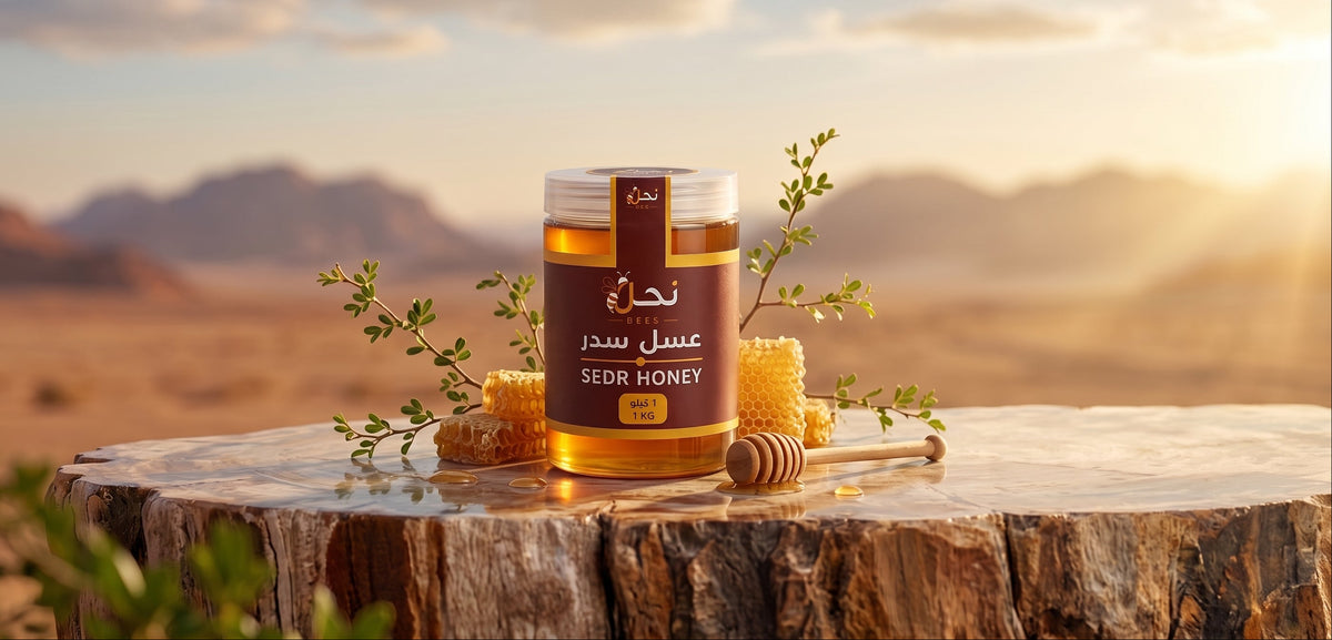 A jar of honey with Arabic and English labels sits on a rustic tree stump, surrounded by honeycomb pieces, a honey dipper, and small green sprigs, with a blurred desert landscape at sunset in the background.